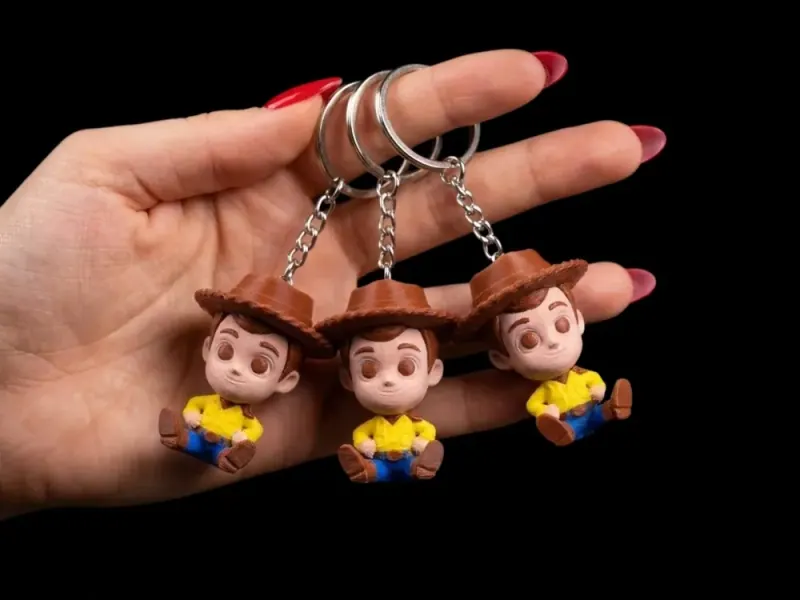 Três chaveiros Woody suspensos pelas argolas na mão, com chapéu e olhos marrons impressos, camisa amarela e calça azul; textura de filamento aparente