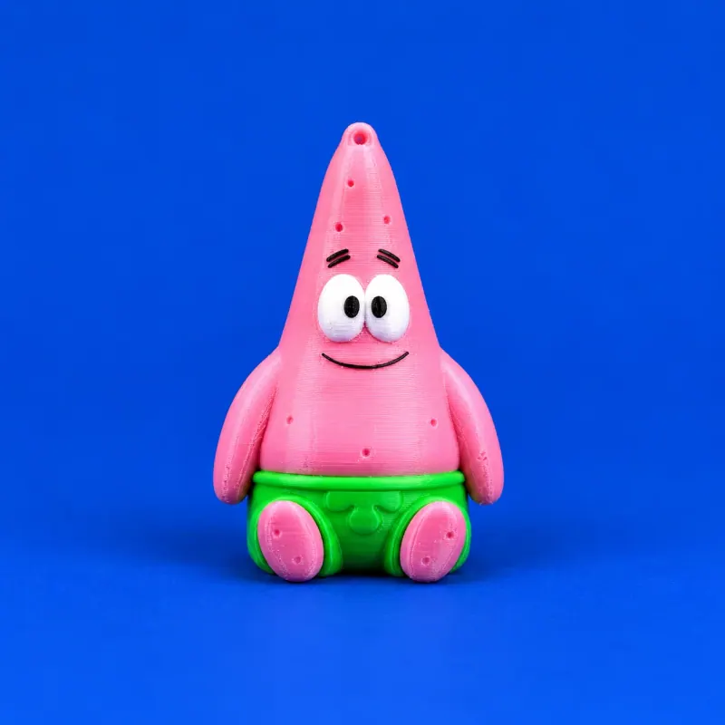 Uma miniatura Patrick rosa sentada em fundo azul, com shorts verdes, olhos brancos e pretos impressos e furo integrado no topo