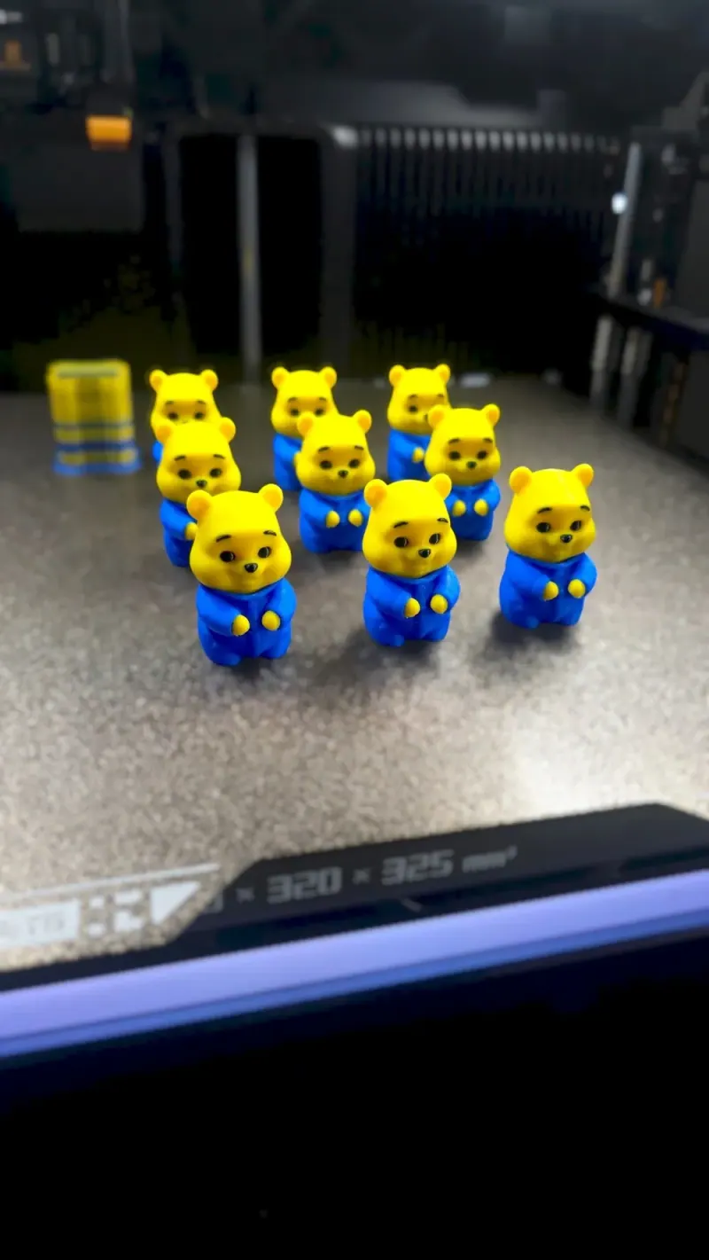 Lote de Baby Pooh amarelo com roupa azul e pequenos olhos, sobrancelhas e nariz pretos, apoiado na mesa de impressão