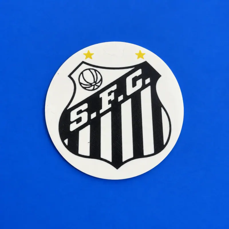 Porta-copo circular Santos branco e preto com duas estrelas amarelas, deitado sobre fundo azul