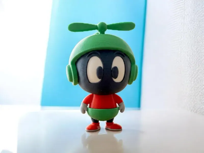 Marvin bebê com capacete verde e adereço horizontal no topo, rosto preto e olhos claros impressos, roupa vermelha e verde e sapatos vermelhos apoiados sobre mesa