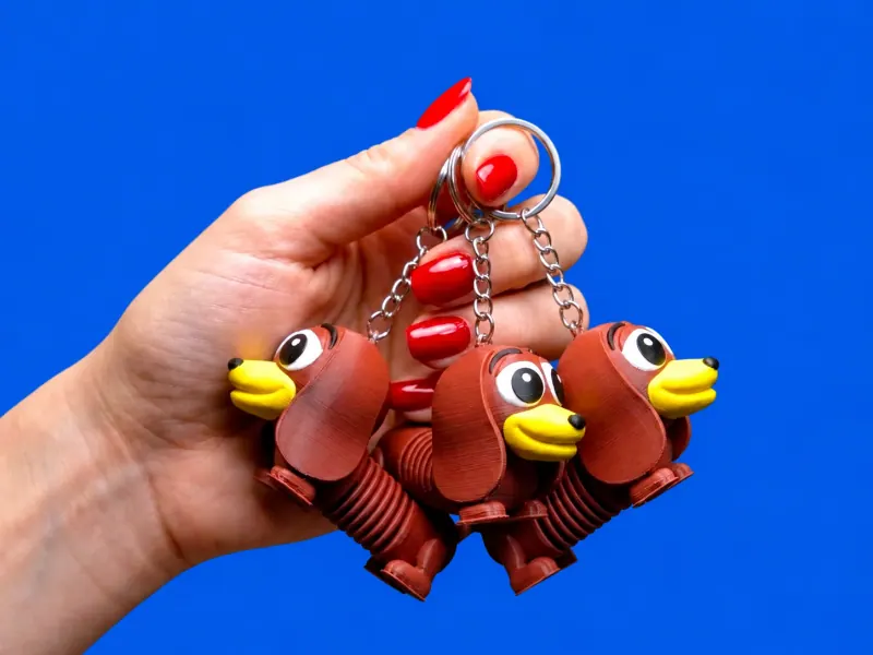 Três chaveiros Slinky marrons com focinho amarelo, olhos brancos e pretos impressos e corpo articulado de filamento, sustentados pelas correntes e argolas na mão