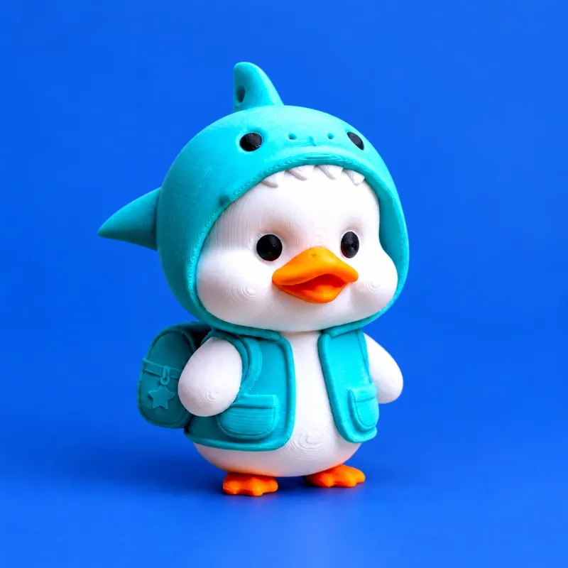 Patinho branco de bico e pés laranja usando capuz de tubarão turquesa com dentes, nadadeiras e mochilinha, com olhos pretos impressos e pés apoiados sobre fundo azul