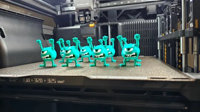 Lote de Mike Wazowski turquesa com braços levantados, olho único branco e preto e dentes claros, apoiado pelos pés na mesa de impressão